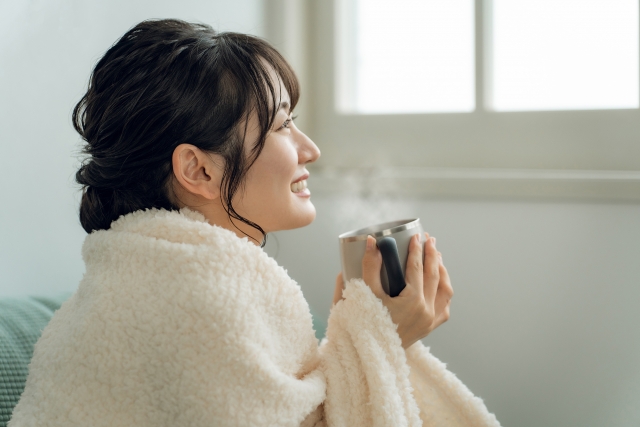 毛布をかぶり温かい飲み物で冷えた体を温める女性